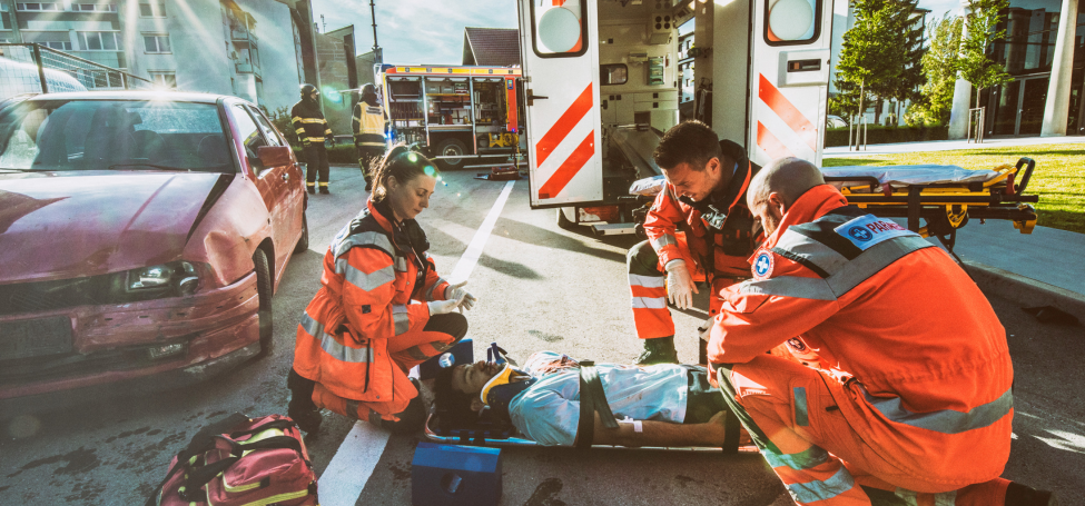 Paramedics tending to an injured person at a vehicle accident scene with emergency vehicles in the background.