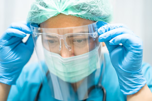A medical professional wearing a clear protective face shield, surgical cap, mask, and blue nitrile gloves to prevent eye injuries from splashes and chemicals in the workplace