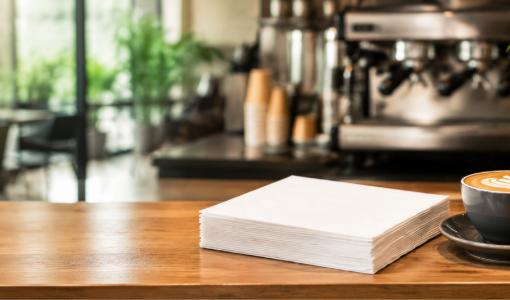 A clean café counter with neatly folded white lunch napkins beside a coffee cup and cutlery